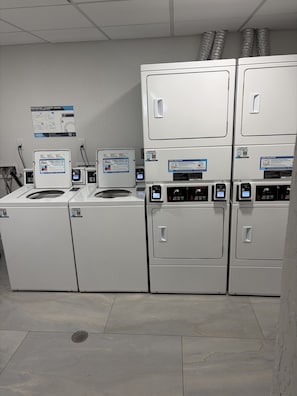 Eine Wäschemöglichkeit mit mehreren Waschmaschinen und Trocknern, einem Schwarzesbrett mit Informationen rund um die Wäsche und einem Lüftungssystem an der Decke.