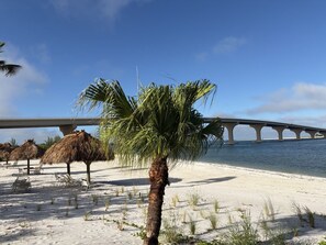 Uma praia com palmeira, guarda-sóis de palha e uma ponte ao fundo.