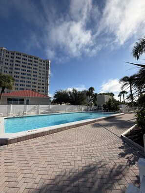A swimming pool with a brick deck, surrounded by palm trees and a tall hotel building in the background. - One Bedroom Condo with Water View #601 (Fort Myers Beach)