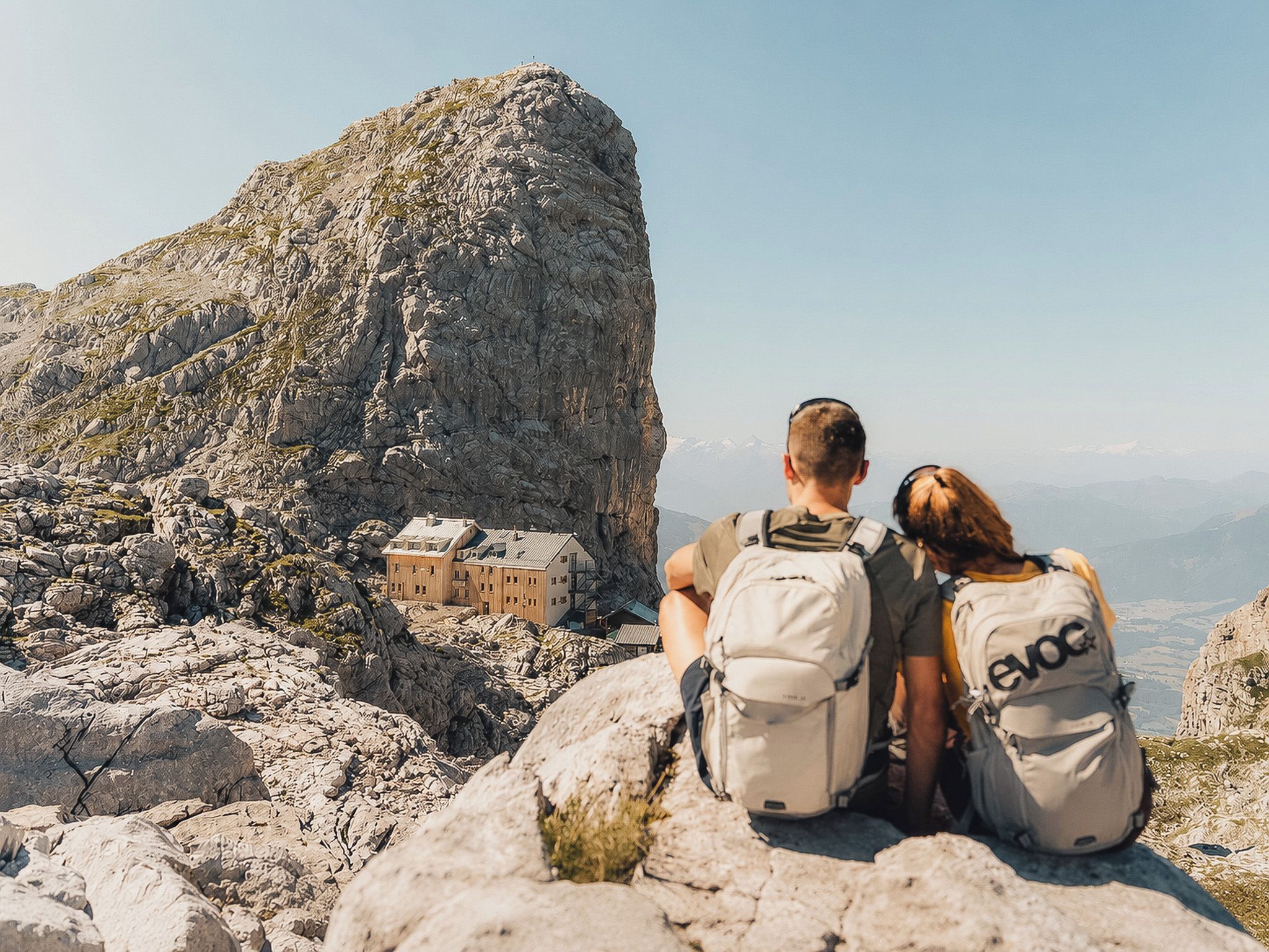 Two people sitting on a rocky outcrop overlooking a mountainous landscape with a small building in the distance.