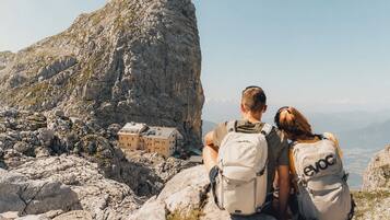 Two people sitting on a rocky outcrop overlooking a mountainous landscape with a small building in the distance.
