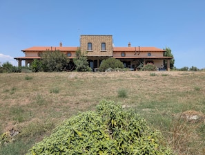 Un edificio grande de dos pisos con techo de tejas rojas, rodeado por un campo cubierto de césped y vegetación.