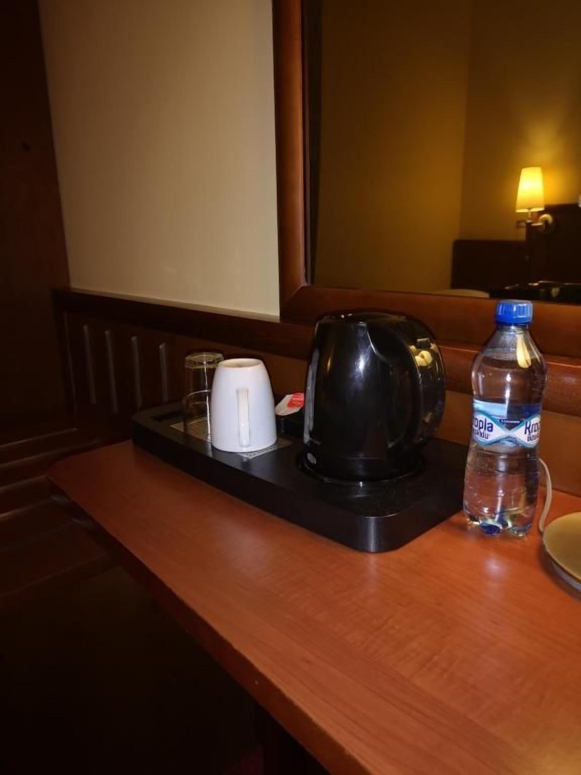 A hotel room with a wooden table, a black electric kettle, a white cup, a bottle of water, and a tray.