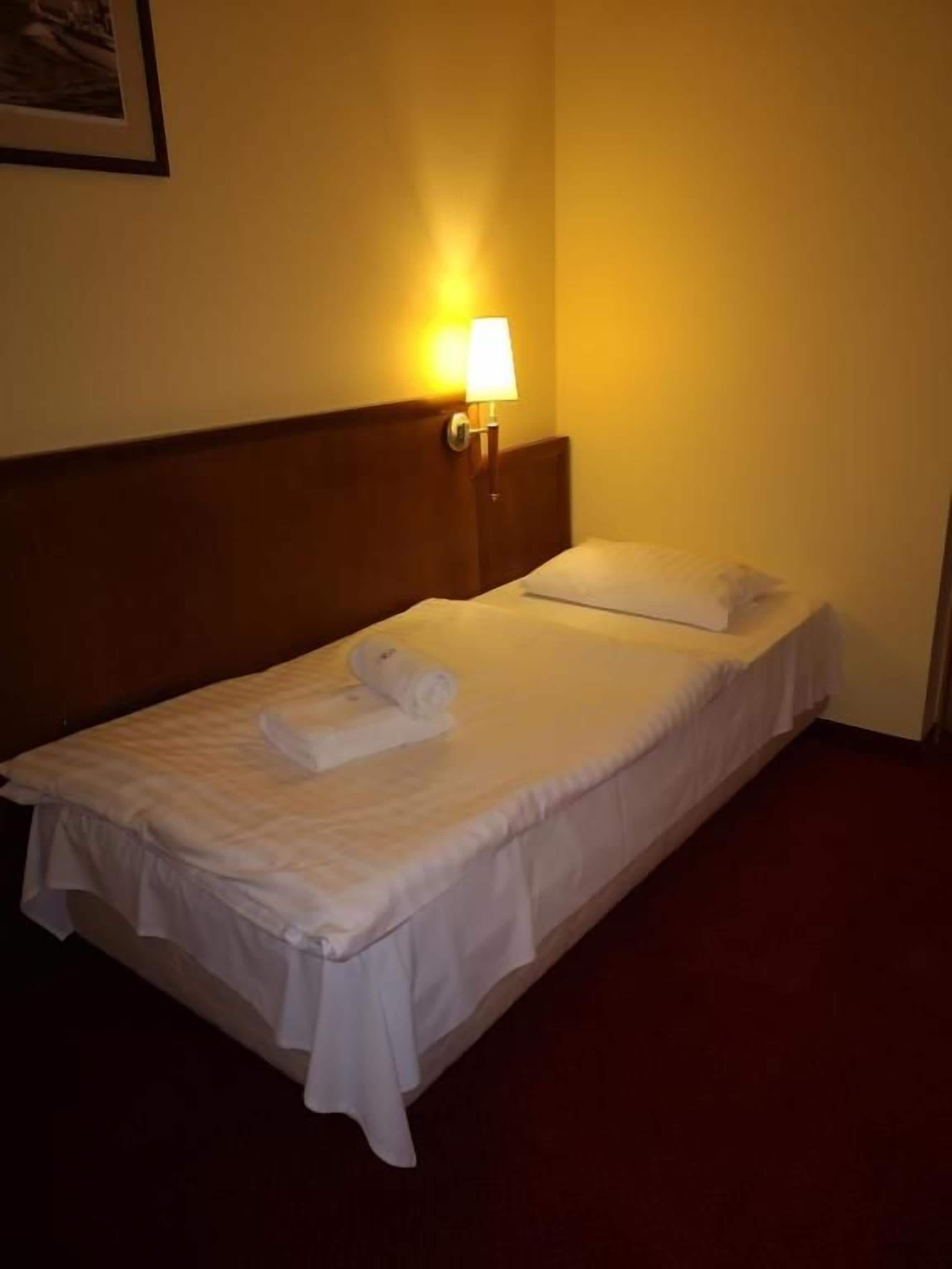 Un lit simple avec une tête de lit, une lampe fixée au mur et une serviette enroulée posée sur le lit.