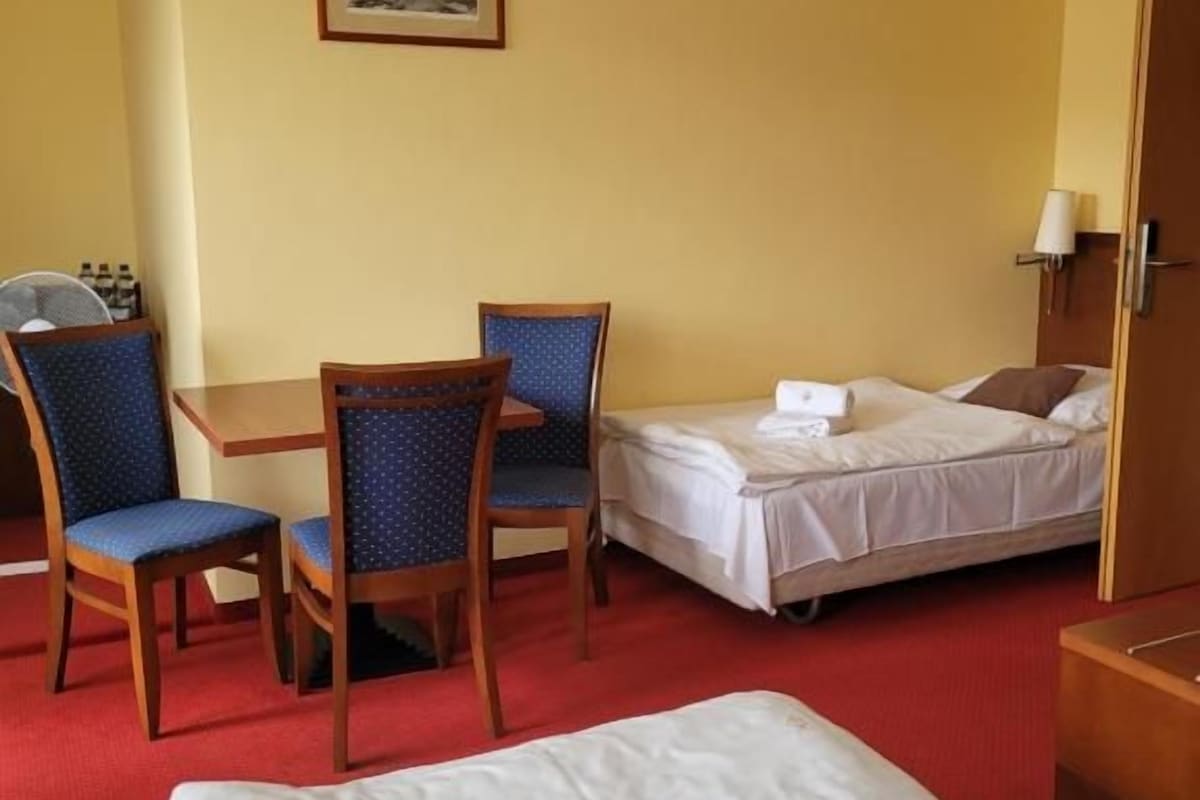 A hotel room with a bed, a desk with two chairs, a bedside table, and a wall-mounted lamp.