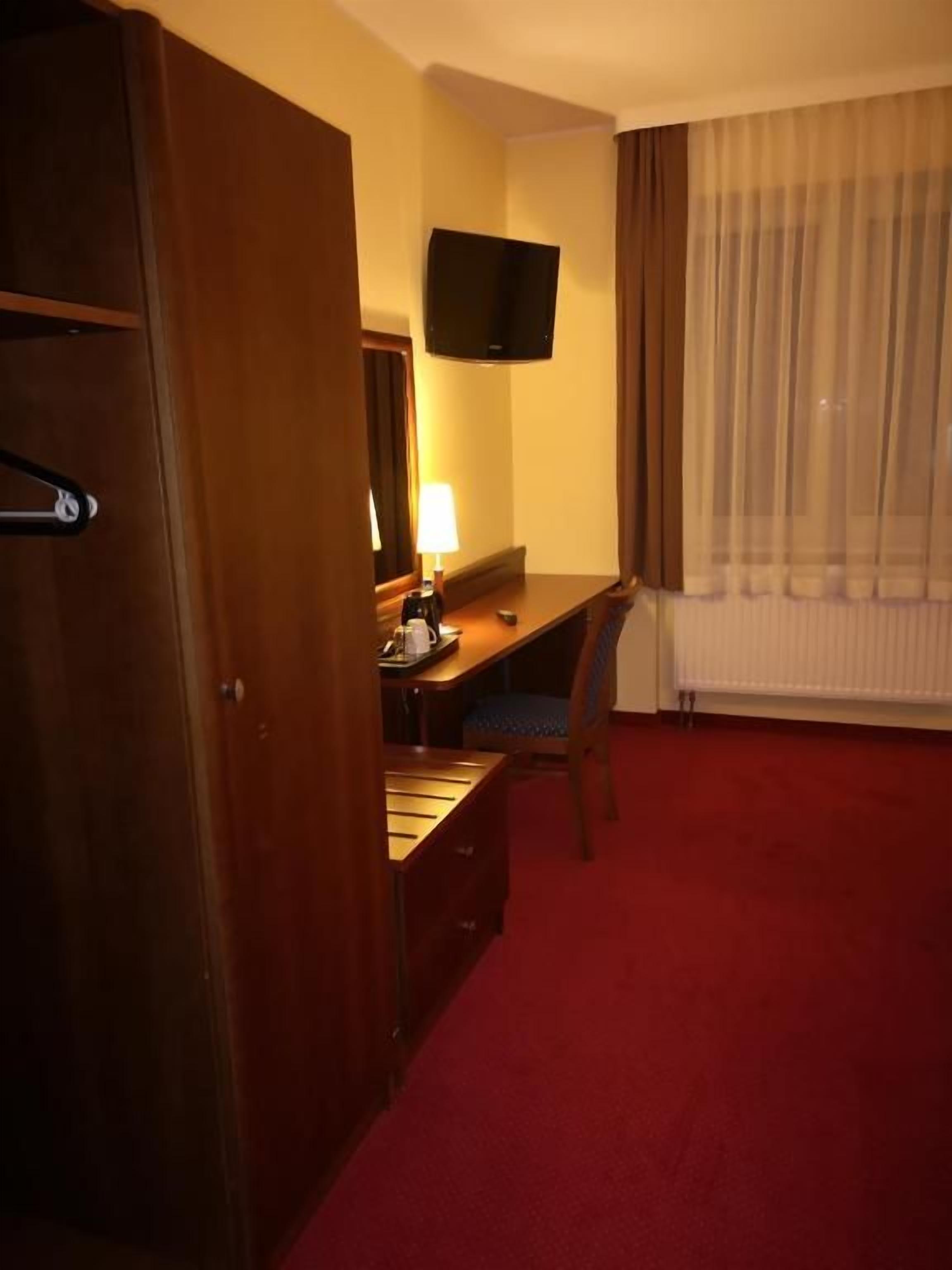 Ett hotellrum med en garderob i trä, ett skrivbord med en lampa, en tv på väggen och en röd matta.
