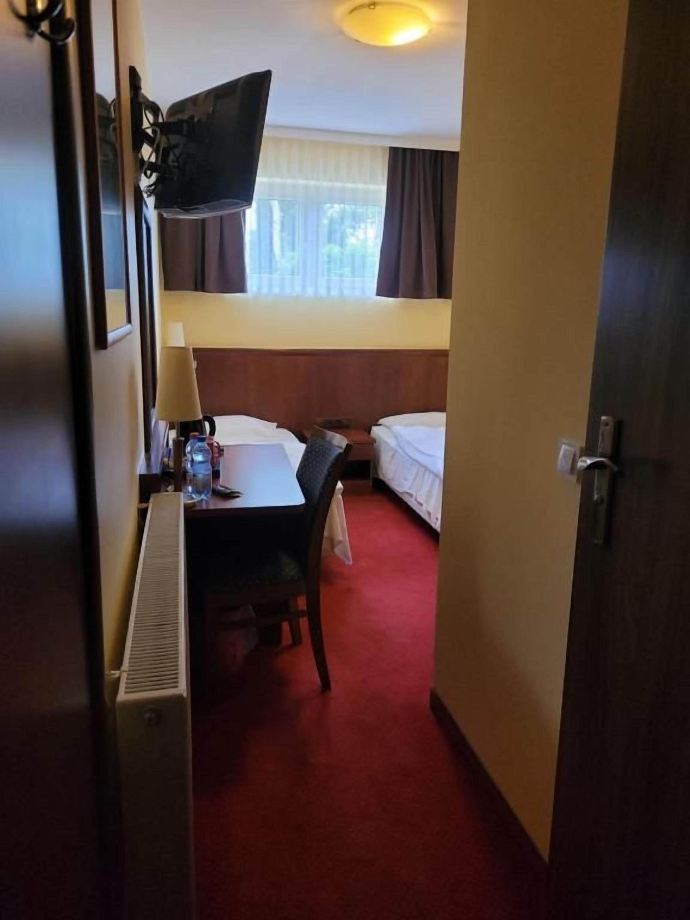 Una habitación de hotel con cama, escritorio con silla, televisor montado en la pared y ventana con cortinas.