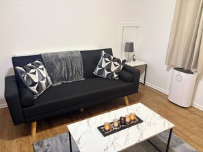 Eine schwarze Couch mit geometrisch gemusterten Kissen, ein Couchtisch aus Marmor mit Kerzen, ein kleiner Nachttisch mit Lampe und ein weißer Heizkörper.