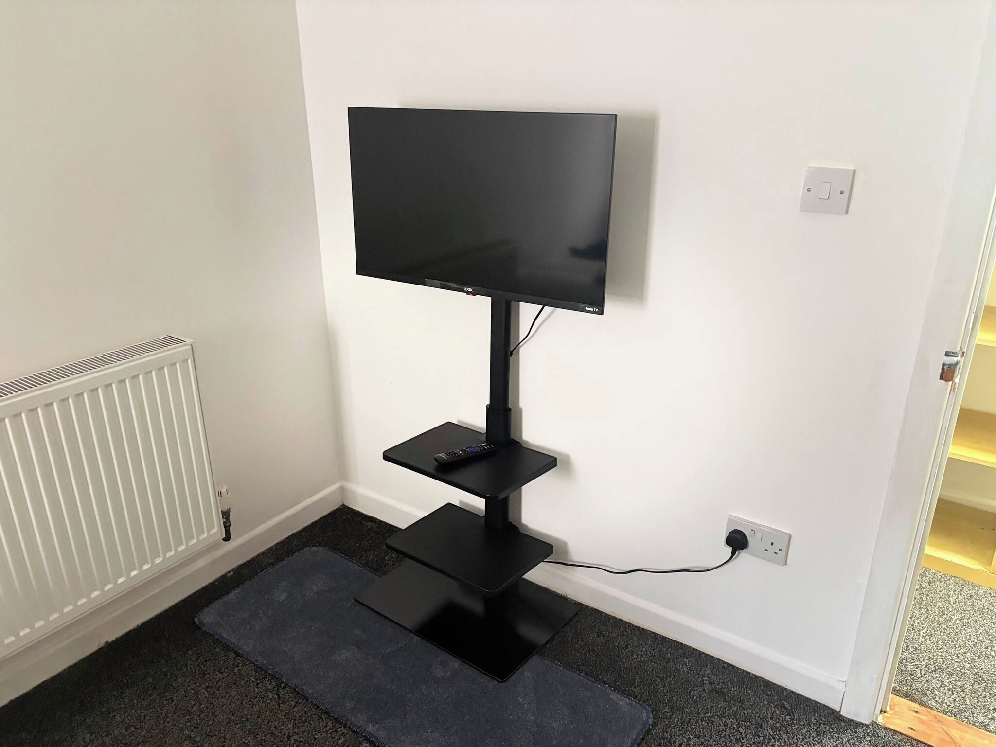 A black TV stand with a flat-screen TV, a remote control, and a power cord plugged into a wall socket.