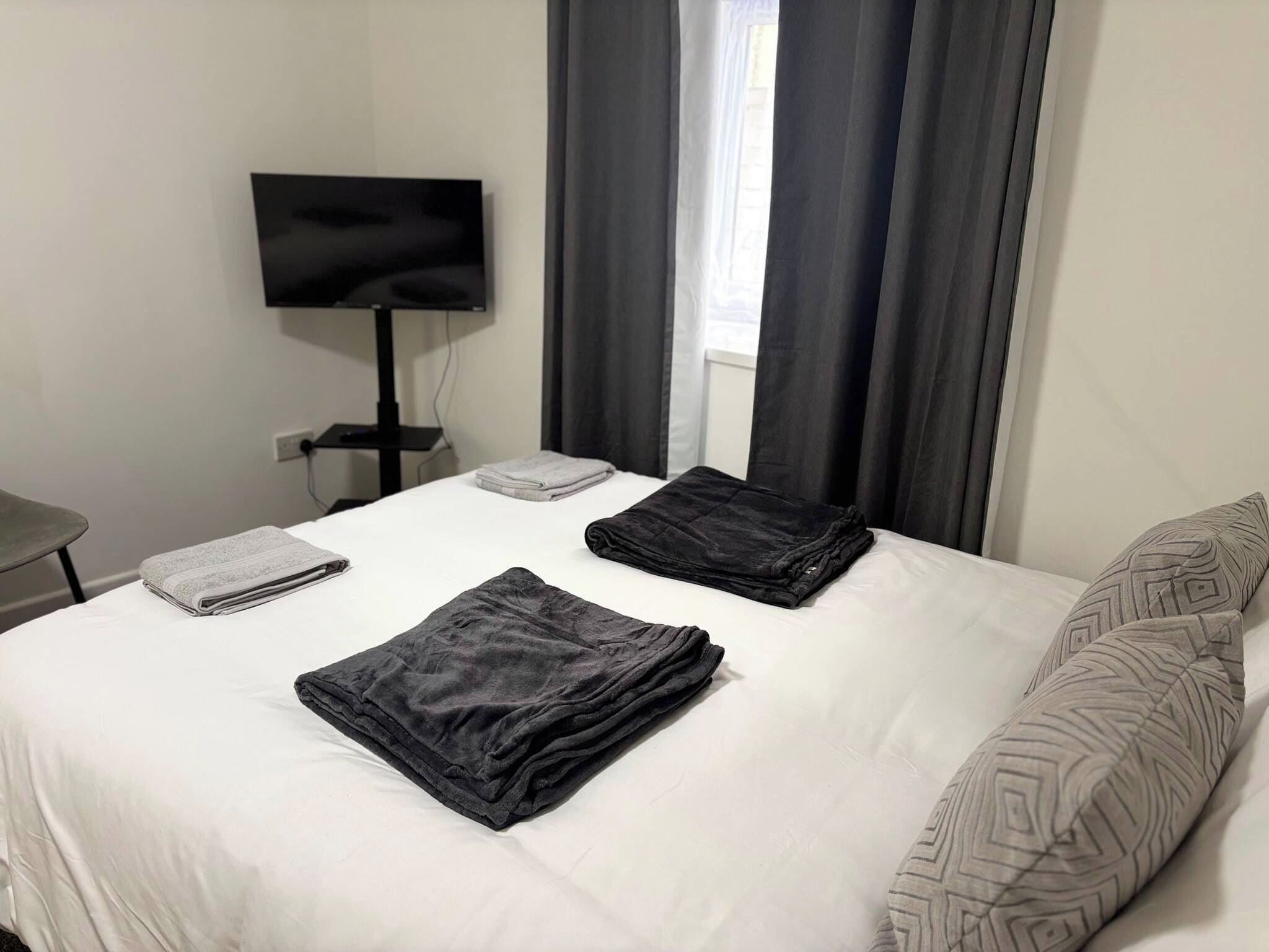 A neatly made bed with folded blankets, a flat-screen TV on a stand, and a window with curtains.