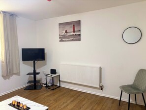 Une pièce avec une télévision, un radiateur, une chaise, une table avec des bougies et un tableau représentant un phare.