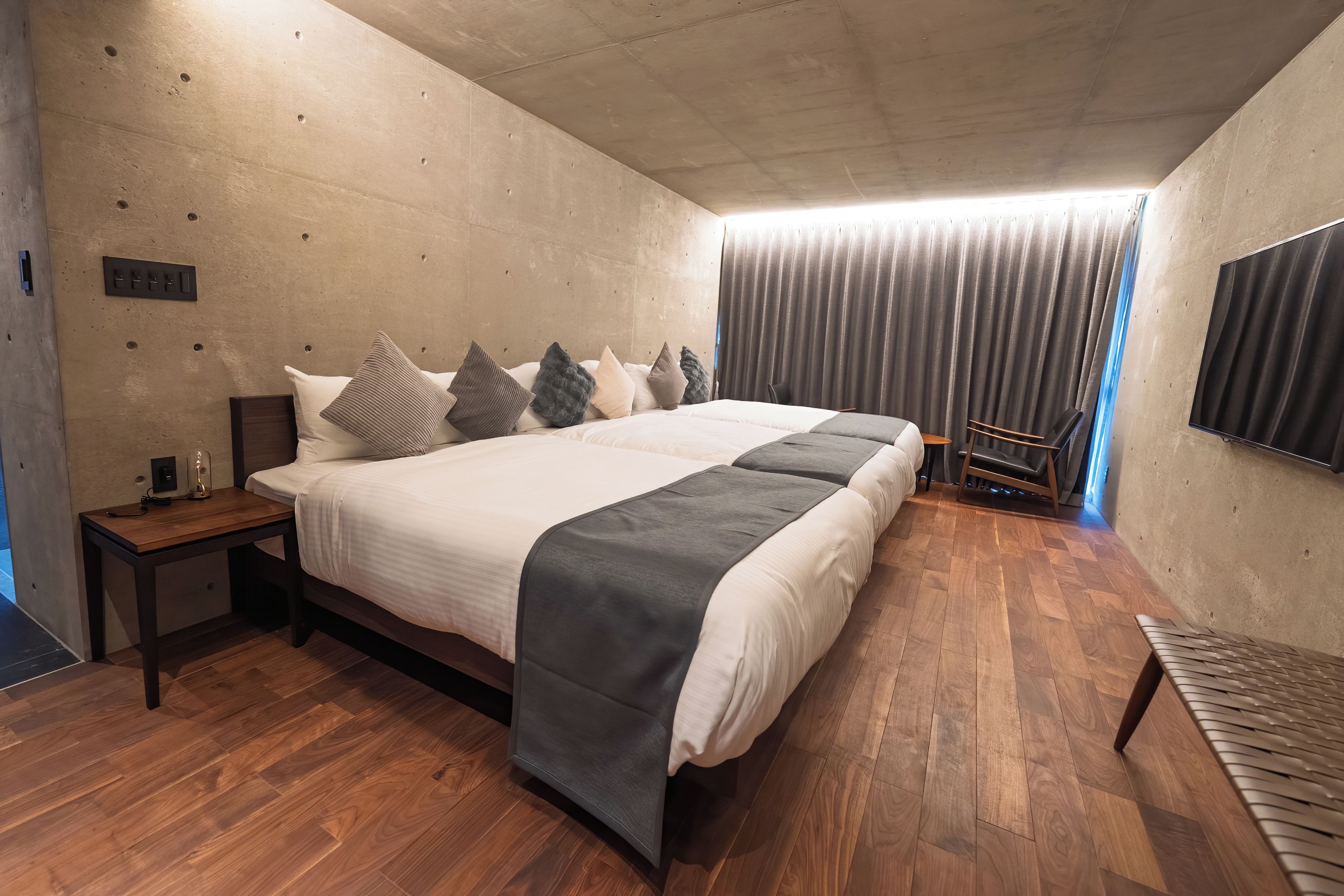 Quarto moderno com uma cama grande, mesa de cabeceira, cadeira, televisão e janela com cortinas.