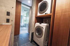 A compact laundry room with a front-loading washer and dryer, wooden shelving, and a tiled floor. - CASA MIYAKO II PREMIUM (Miyakojima)