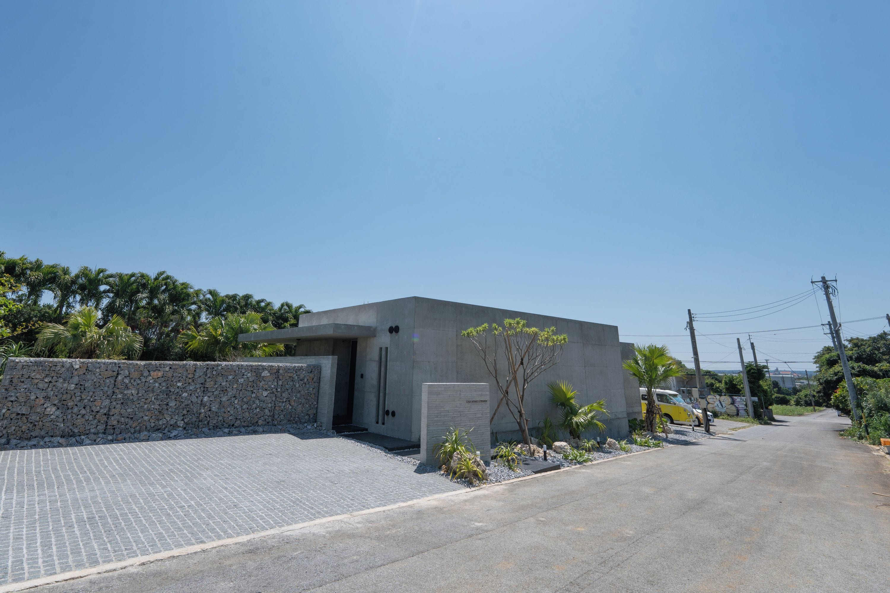 Ein modernes Gebäude mit Betonfassade, einem gepflasterten Auffahrtsweg und einem kleinen Garten mit Baum und Sträuchern.