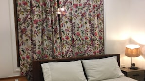 Un lit avec une literie blanche, une table de chevet avec une lampe et une fenêtre ornée de rideaux à motifs floraux.
