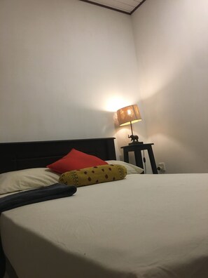 Un lit avec un matelas blanc, un oreiller rouge et une couverture jaune à motifs rouges. Une lampe se trouve sur une table de chevet, à côté du lit.
