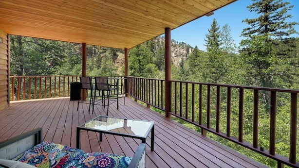 A covered deck with a glass table, chairs, and a sofa, overlooking a forested area. - Margaret Bridge Suite at Rim Rock Lodge (Spearfish)