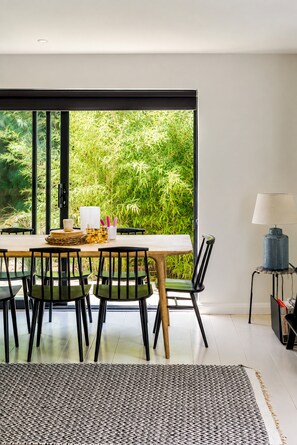 A dining area with a table set for four, chairs, a lamp, and a view of greenery through large windows. - Palm Beach House | 5 Bed by Pett Level Beach - Pass the Keys (Hastings)