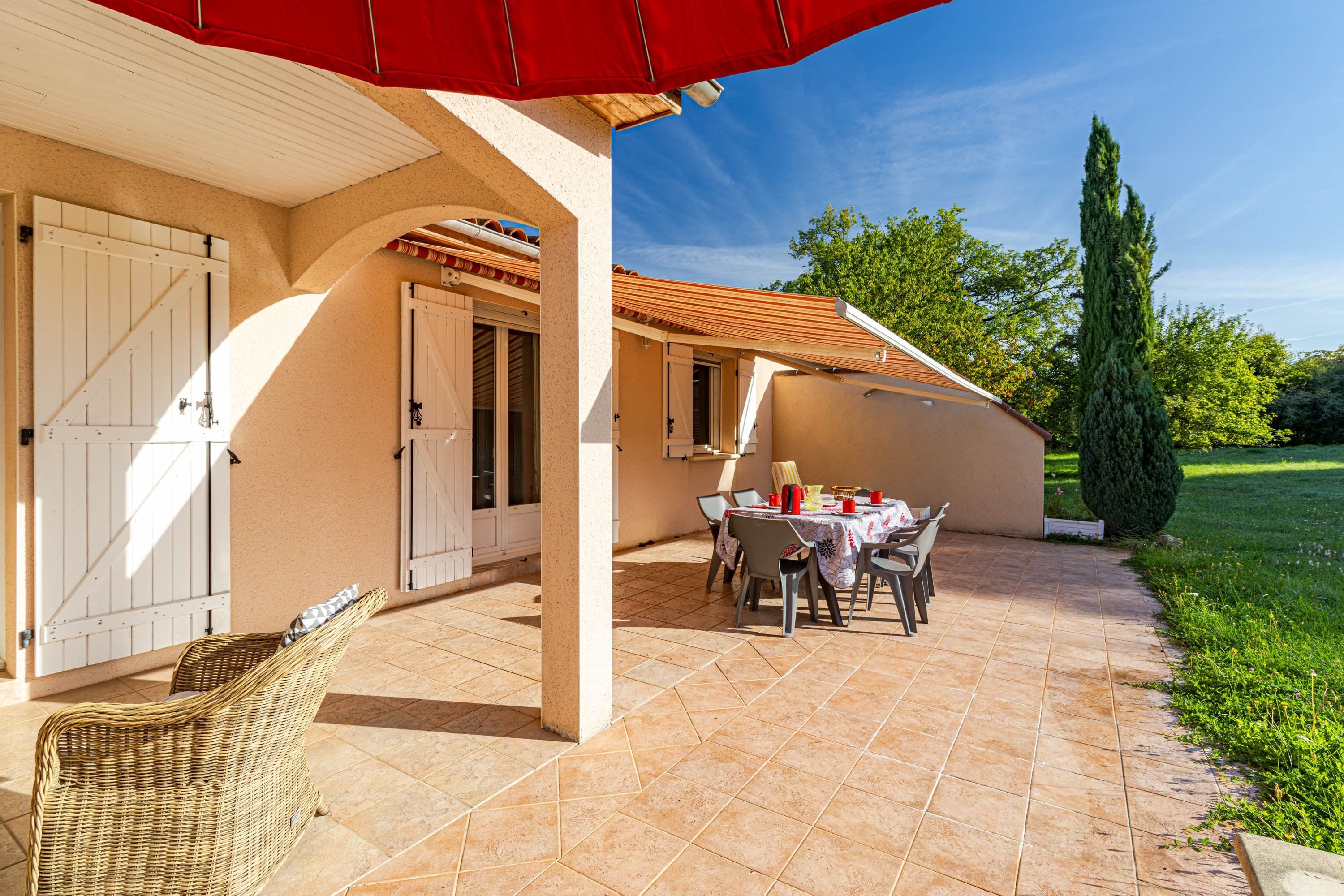 Un patio couvert avec une table dressée pour le repas, une chaise en osier et un parasol rouge.