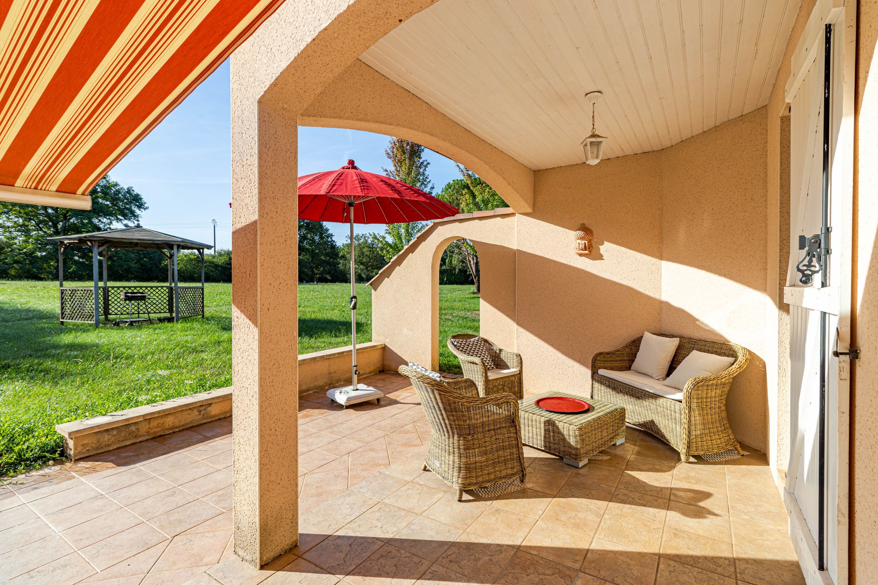 Un espace extérieur abrité, aménagé avec des meubles en osier et agrémenté d’un parasol rouge.