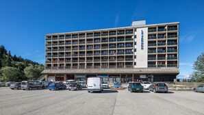 Ein mehrstöckiges Hotel mit zentralem Eingang, einem Parkplatz mit mehreren Autos und einer Bergkulisse.