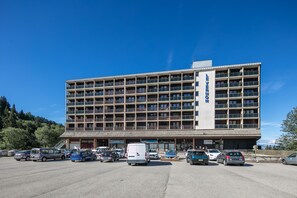 Ein mehrstöckiges Hotel mit zentralem Eingang, einem Parkplatz mit mehreren Autos und einer Bergkulisse.