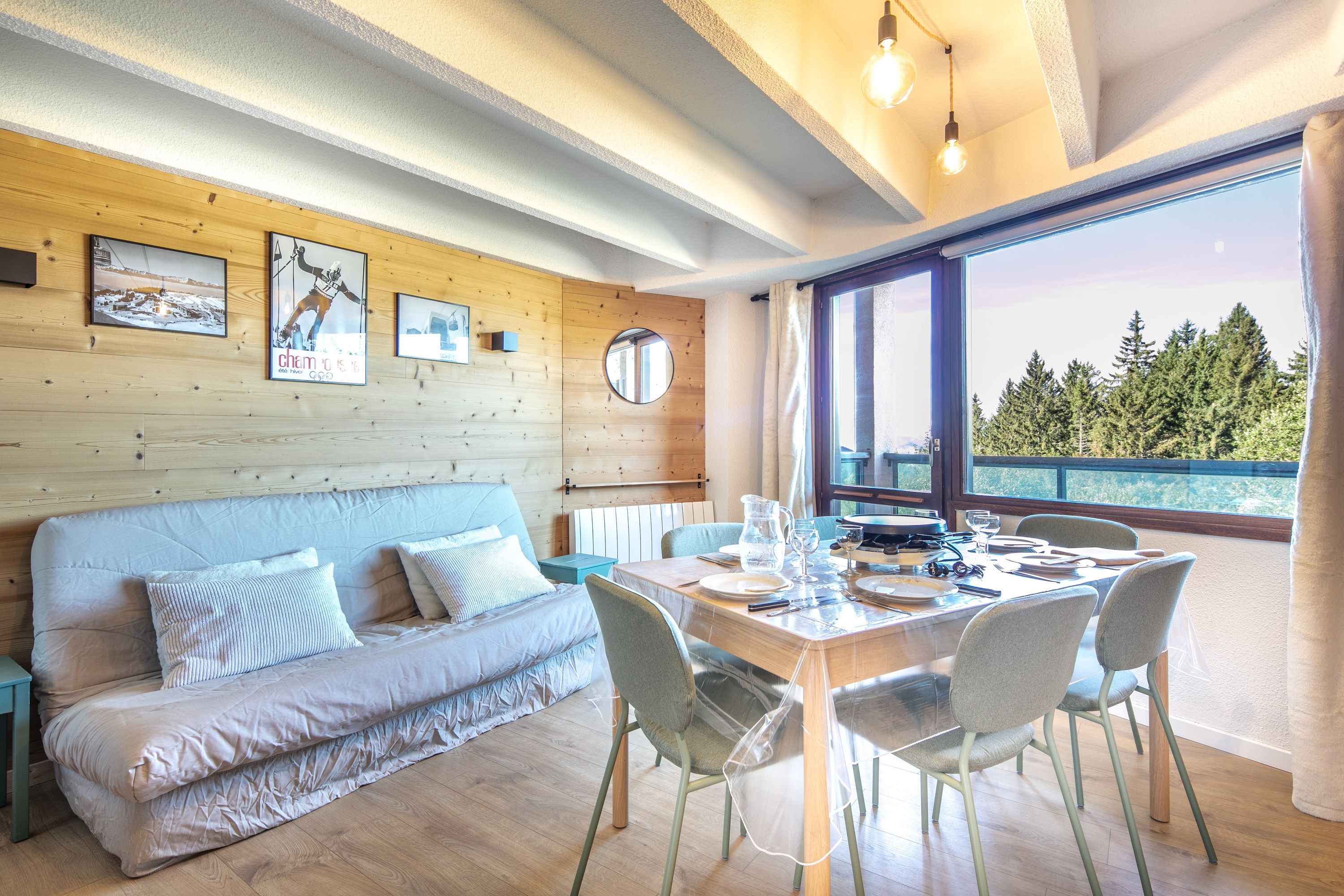 A modern room with a wooden ceiling, a wooden wall, a sofa, a dining table set for two, and a large window with a view of trees.