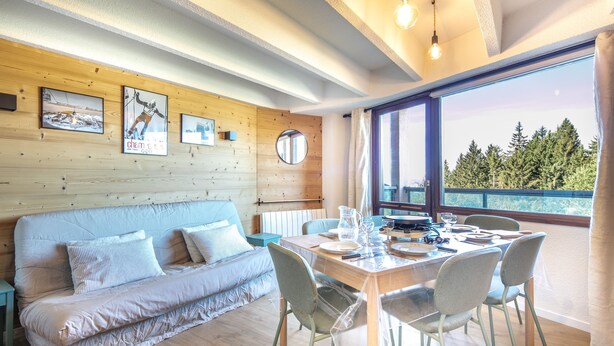 Ein modernes Zimmer mit Holzboden, einer Holzwand, einer Couch, einem Esstisch für zwei Personen und einem großen Fenster mit Blick auf Bäume.