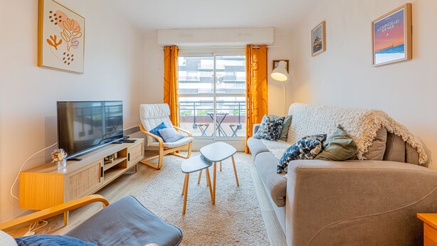 Een moderne woonkamer met een flatscreen televisie, een bank, een stoel, een bijzettafeltje en een balkon met uitzicht op gebouwen.