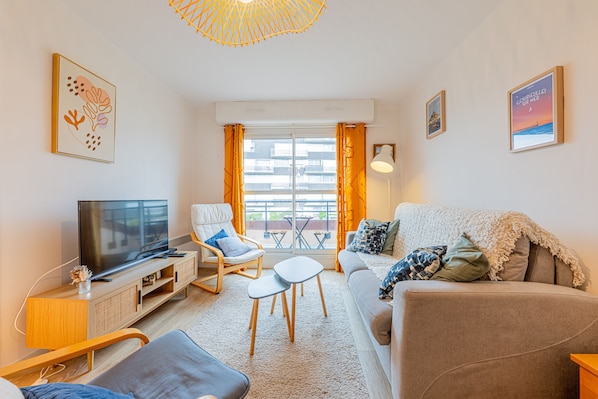 Ein modernes Wohnzimmer mit einem Flachbildfernseher, einem Sofa, einem Sessel, einem kleinen Tisch und einem Balkon mit Blick auf Gebäude.