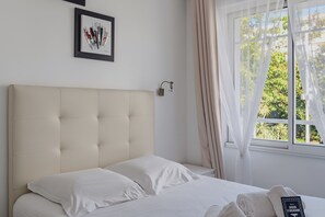 A bedroom with a large bed, a headboard, a bedside table with a lamp, a window with curtains, and a framed picture on the wall. - ArcachonAir plein centre dArcachon à 2 pas de la plage (Arcachon)