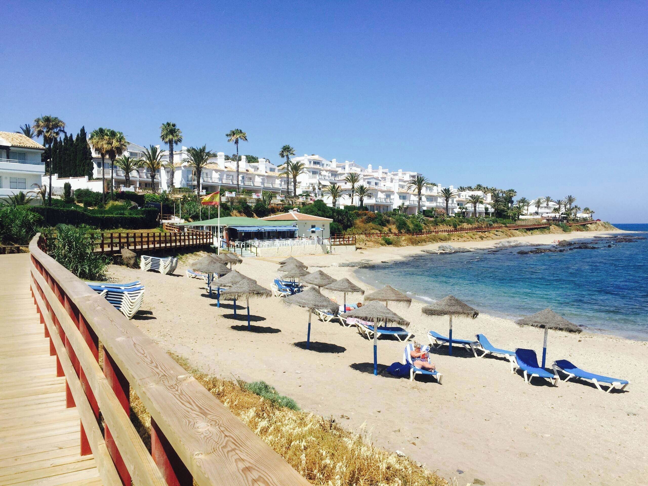 A beach with sun loungers and umbrellas, a wooden boardwalk, and buildings in the background.