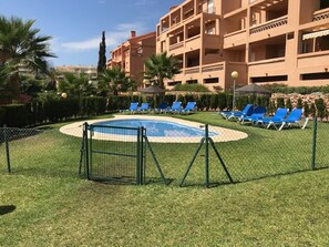 Ein Swimmingpool, umgeben von einem Zaun, mit Liegestühlen und einem mehrstöckigen Gebäude im Hintergrund.