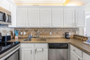A modern kitchen with white cabinets, a granite countertop, a stainless steel sink, and a coffee maker. - Romar Place 1105 (Orange Beach)