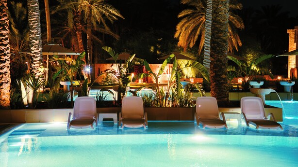Un espace aménagé au bord de la piscine, avec des chaises longues et des palmiers, le tout illuminé la nuit.