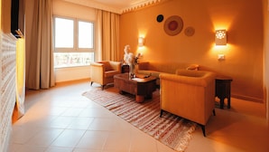 Ein Wohnzimmer mit einer Couch, Sesseln, einem Couchtisch und einem Teppich. Ein Fenster mit Vorhängen, Wandleuchten und einem dekorativen Wandbild.