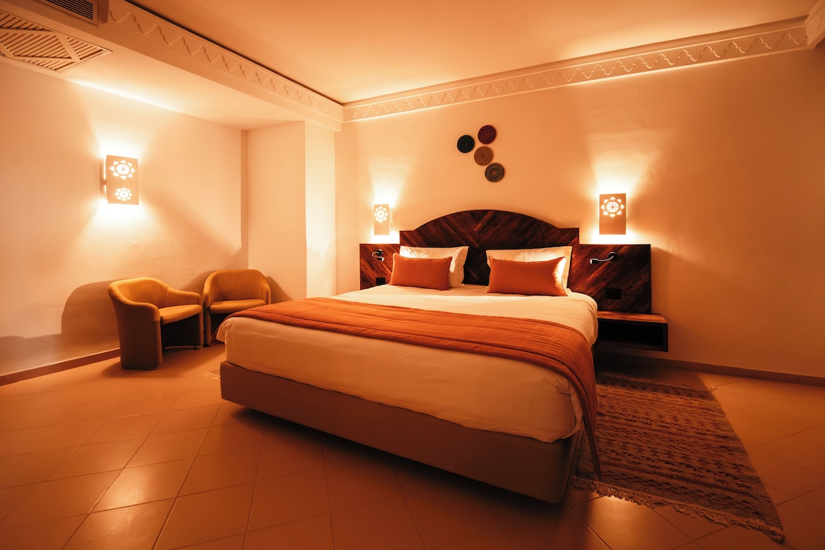Un dormitorio con una cama grande, dos mesitas de noche, una silla y apliques decorativos en la pared.