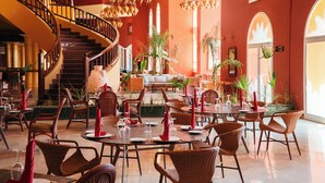 Un vaste espace dédié à la salle à manger, avec de nombreuses tables et des chaises en osier, un magnifique escalier et un grand lustre.