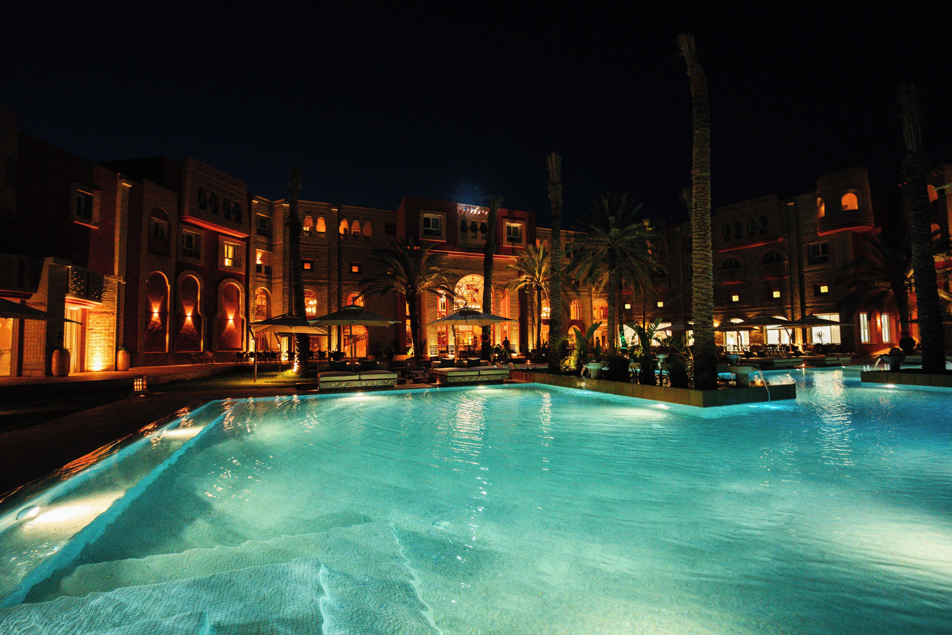 A nighttime view of a hotel with a swimming pool, palm trees, and illuminated buildings.
