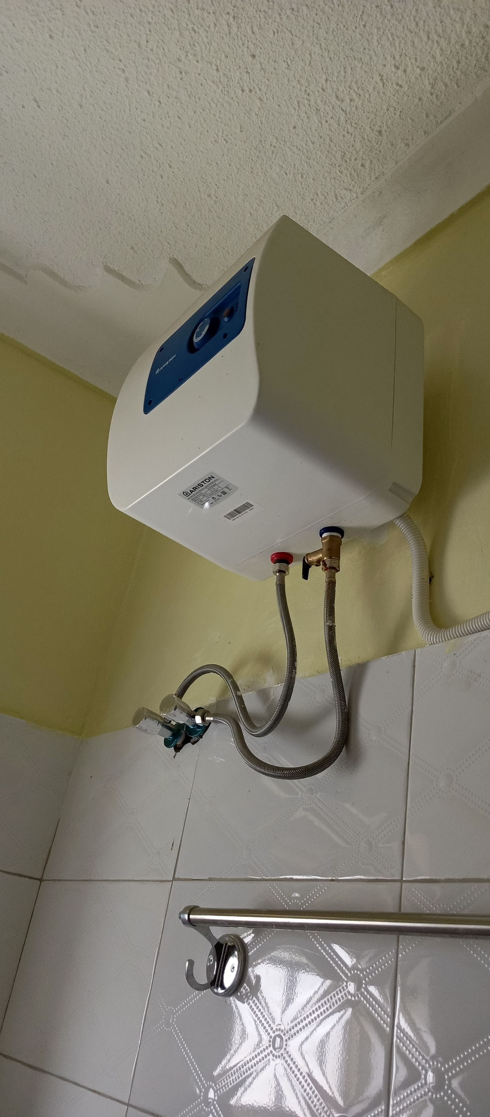 A white water heater mounted on a wall with a blue control panel, connected to a flexible hose and a metal pipe.
