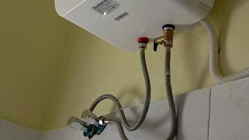 A white water heater mounted on a wall with a blue control panel, connected to a flexible hose and a metal pipe.