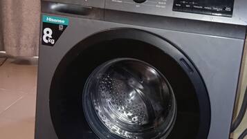 A front-loading Hisense 8kg front-loading washing machine with a control panel, located in a room with a tiled floor and a curtain.