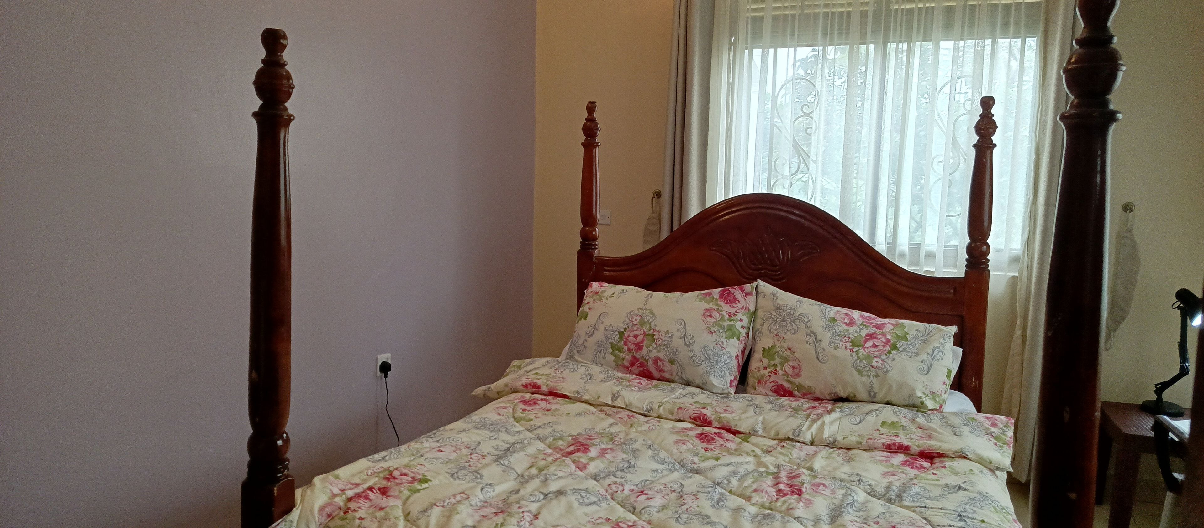 Una cama con dosel, ropa de cama floral, un cabecero de madera y una ventana con cortinas translúcidas.
