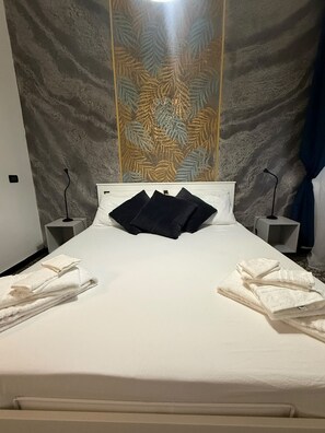 A neatly made bed with white linens and two dark pillows, a bedside table with a lamp, and a decorative wall with leaf patterns. - Spacious 4-bedroom apartment in delightful Genova with AC (Genova)