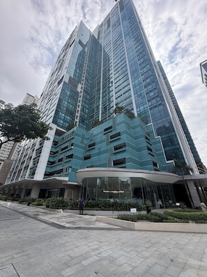 A modern high-rise building with a glass facade, a curved entrance, and a landscaped area with plants. - New! Roomy & Zen Wi-fi 350Mbps  Uptown BGC, One Uptown Residence (Taguig)