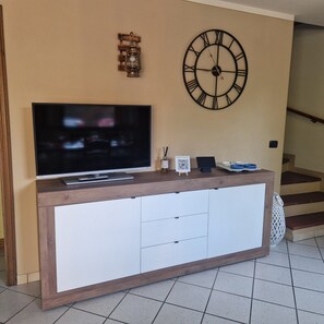 Een moderne woonkamer met een flatscreen televisie op een houten dressoir, een decoratieve wandklok en een klein tafeltje met een QR-code.