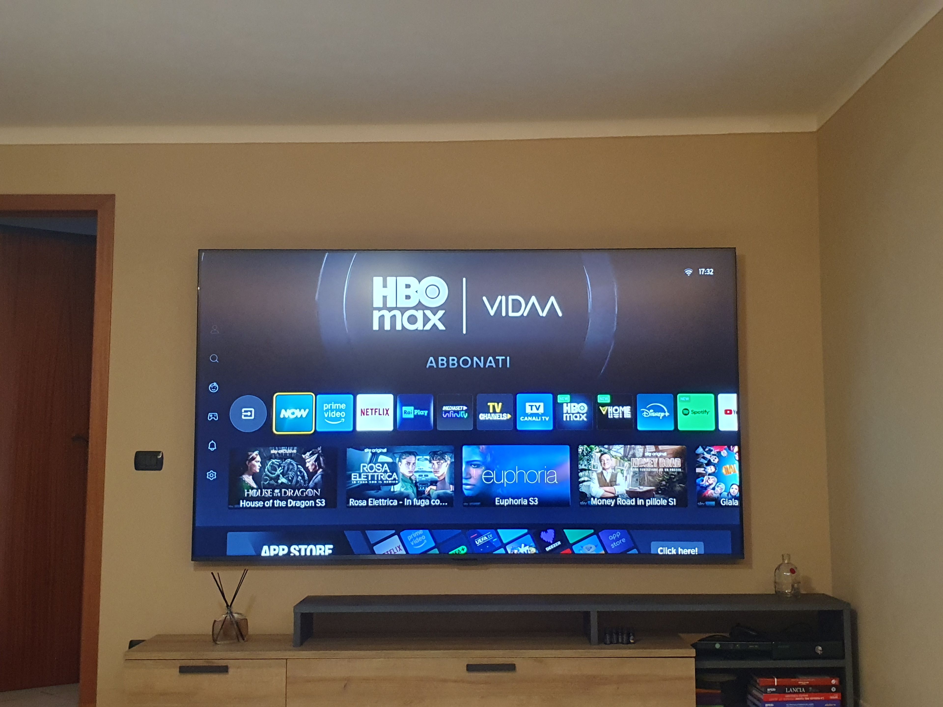 Televisão de tela plana presa na parede exibindo a interface HBO Max VIDMA com diversos serviços de streaming e miniaturas de conteúdo.