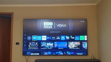 Televisão de tela plana presa na parede exibindo a interface HBO Max VIDMA com diversos serviços de streaming e miniaturas de conteúdo.