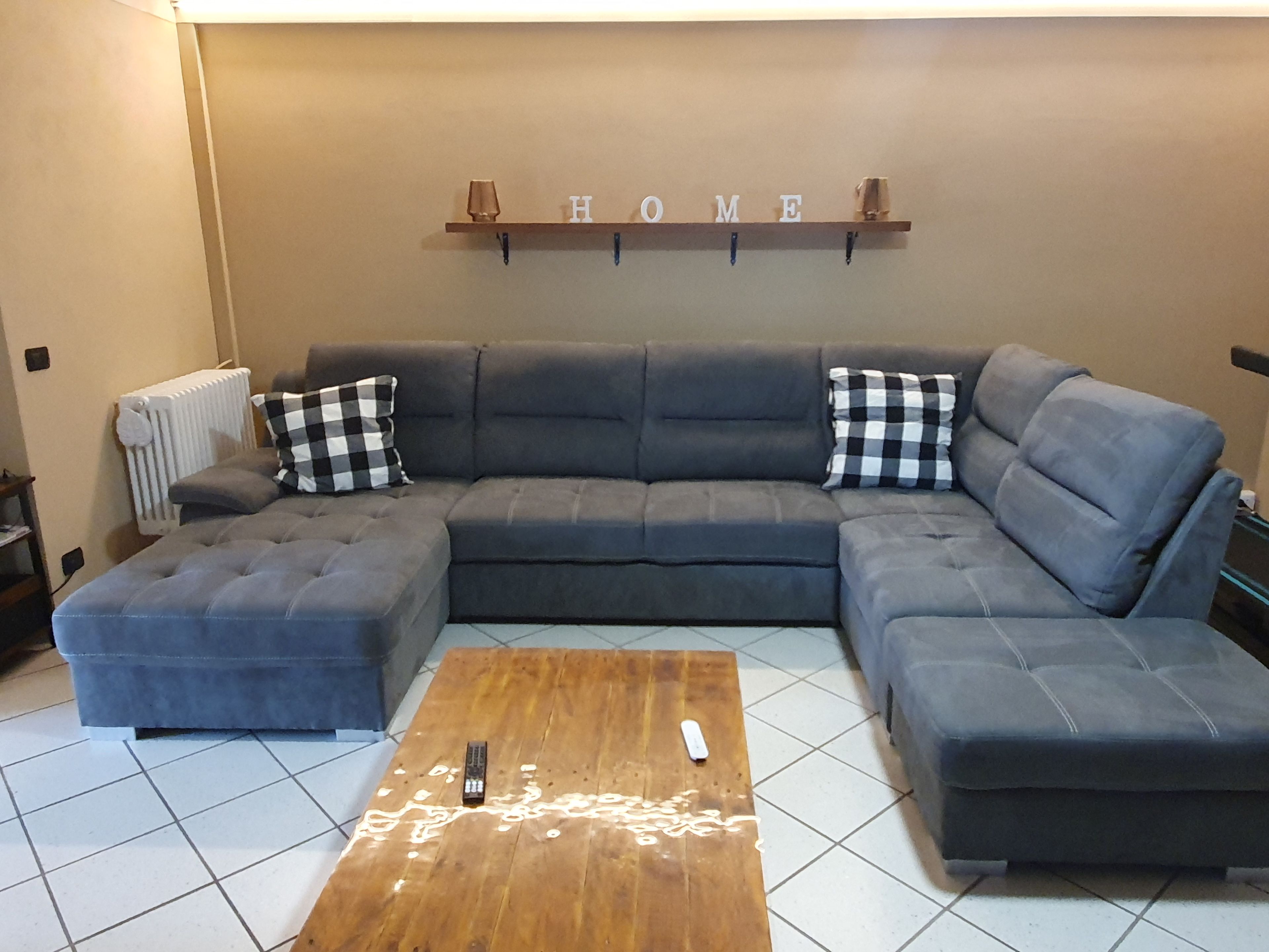 Ein Wohnzimmer mit einem Ecksofa, einem Holztisch und einem an der Wand befestigten Regal mit der Aufschrift ‚HOME‘.