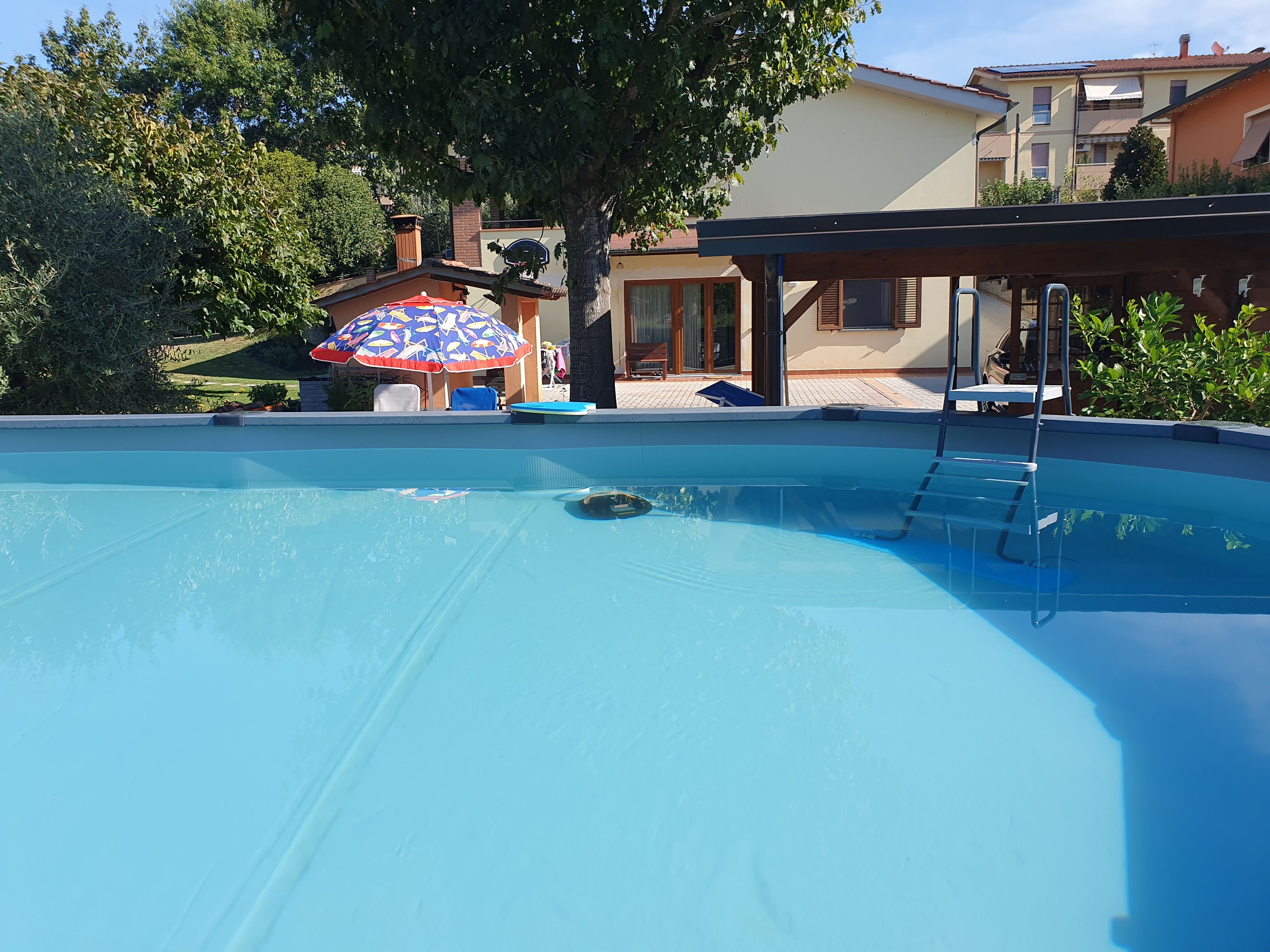 Piscina com escada, área de estar coberta e uma casa ao fundo.